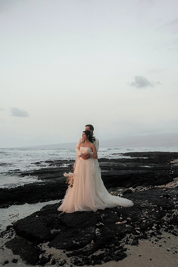 Couple portrait at a rocky beach wedding portrait, groom hugging bride in a strapless dress holding bouquet with ocean waves behind them