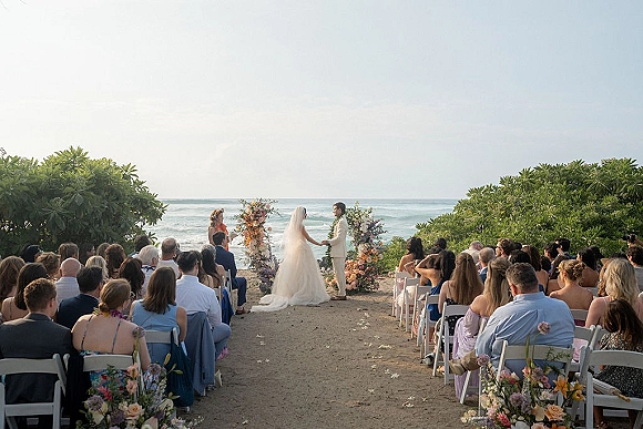 Ceremony moment at an outdoor wedding ceremony with bride and groom holding hands beneath a floral arch by the oceanfront aisle