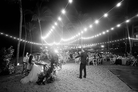 Wedding reception toast as best man gives a speech, bride and groom listen at sweetheart table under string lights on a sandy beach at night