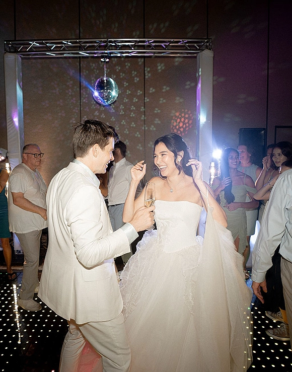 Wedding dance floor first dance reception with bride in strapless gown holding a champagne flute, groom in white tux under disco ball lights