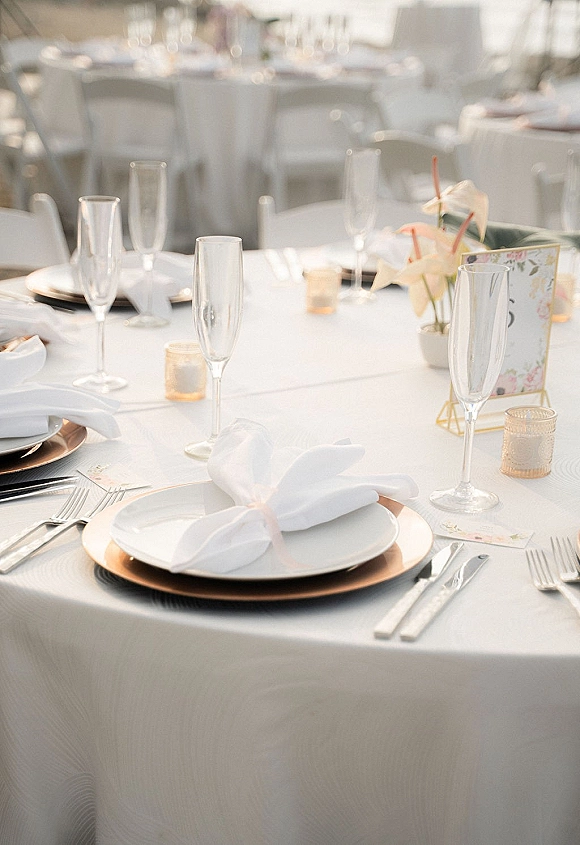Reception tablescape with wedding place setting on white tablecloth, charger plates, silver flatware, champagne flutes, candles, floral centerpiece indoors