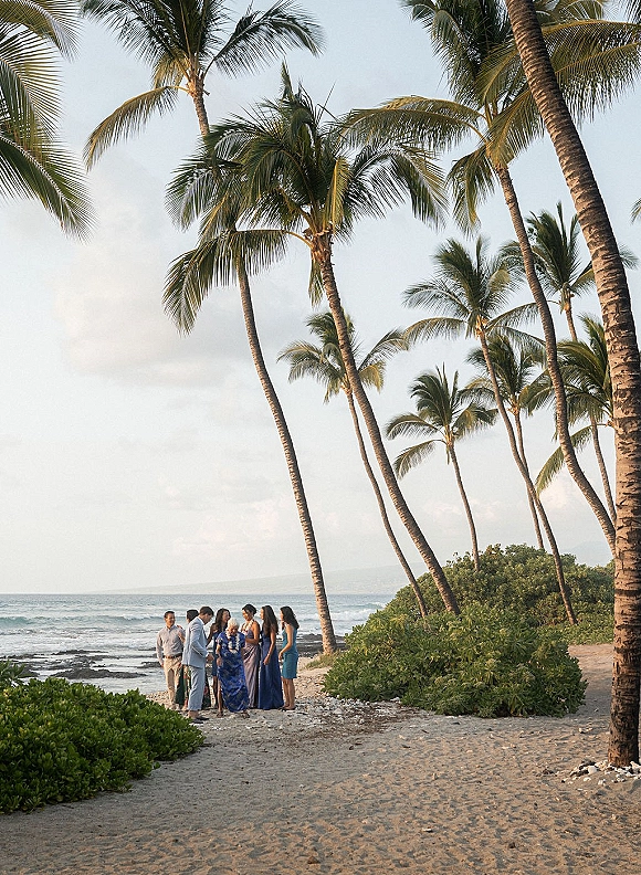 Wedding ceremony on a sandy beach with guests and bridesmaids in blue dresses, ocean and palm trees behind them