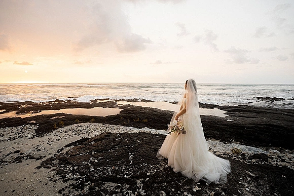 Bridal portrait of a bride on beach holding a bouquet, cathedral veil and dress train blowing in wind beside rocky tide pools at sunset