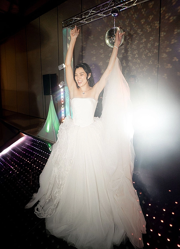Bride dancing on a wedding reception dance floor with arms up, strapless gown and veil glowing under disco ball lights and haze
