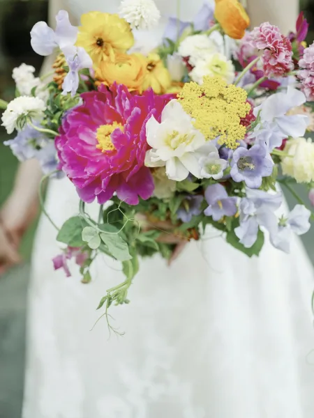 Bridal bouquet with colorful bridal bouquet blooms of peony, ranunculus, and sweet peas with trailing greenery against a white dress outdoors