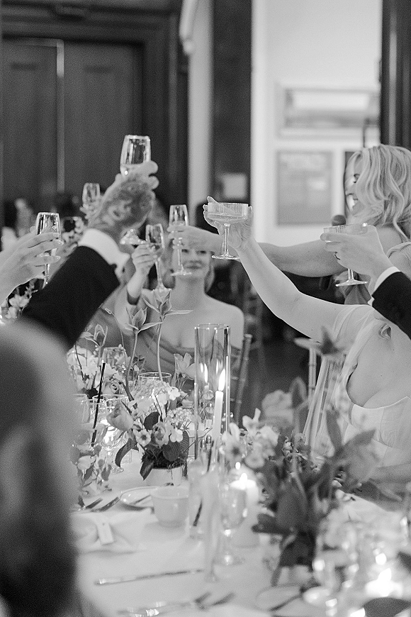 Wedding toast with guests raising champagne flutes over a candlelit reception table with floral centerpieces in an indoor room