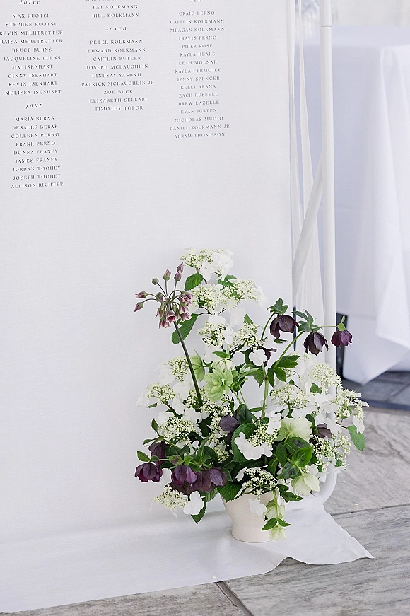 Seating chart sign with wedding seating chart typography, framed by white and dark purple florals in a vase against white tent drape