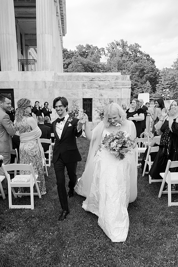 Wedding recessional as bride and groom walk the aisle hand in hand, bouquet and long veil flowing past cheering guests on a lawn by columns
