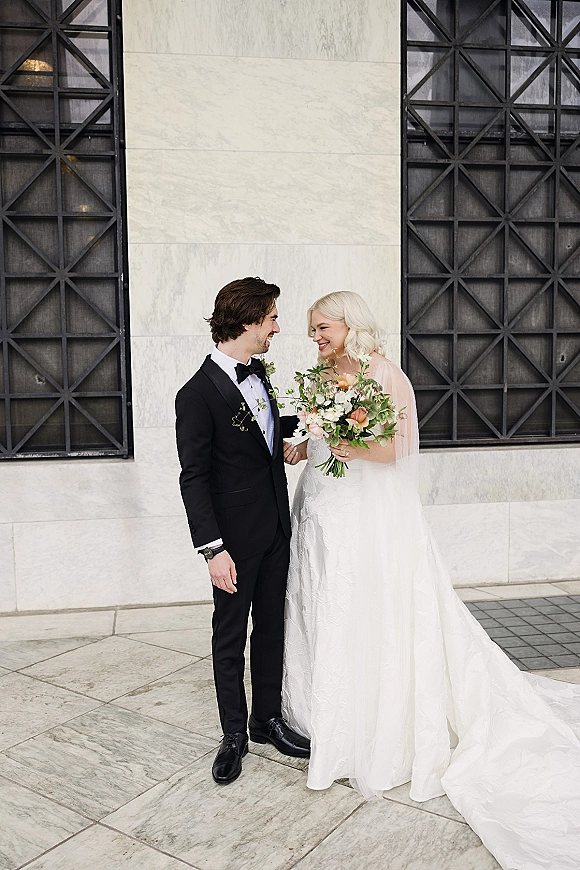 Couple portrait of bride and groom portrait laughing, bride holding bouquet and veil beside groom in black tuxedo against stone wall