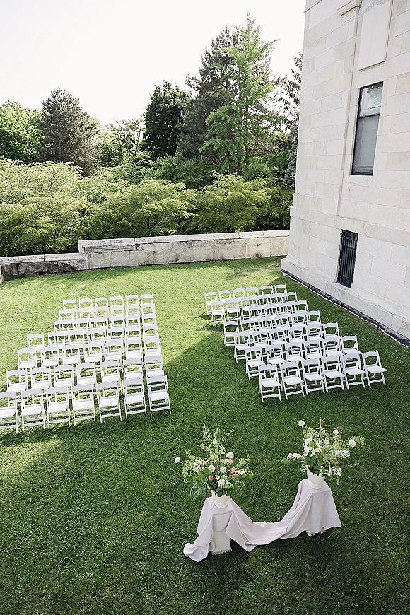 Outdoor ceremony setup with white wedding chair rows on a grass lawn, facing pedestal florals and draped fabric near trees and stone wall