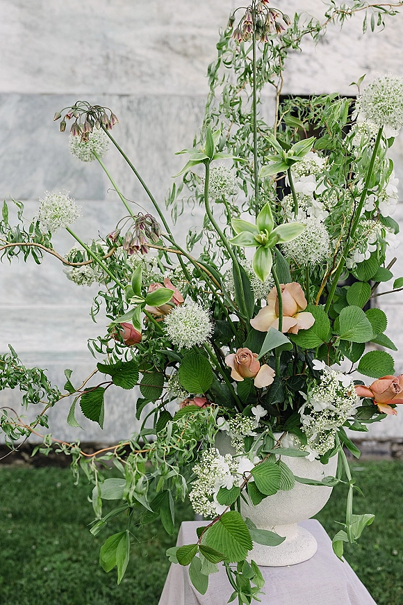 Wedding floral arrangement in a stone urn with peach roses, white allium, hydrangea and trailing greenery on a lawn by a stone wall