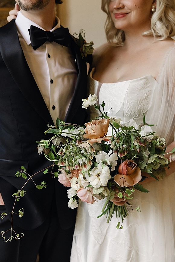 Couple portrait of bride and groom with bride holding a bridal bouquet, veil and strapless dress beside groom in tuxedo against an indoor wall