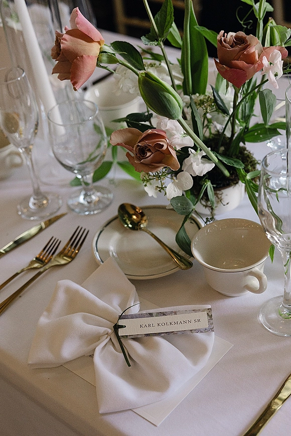 Wedding tablescape with wedding place setting featuring rose and greenery centerpiece, gold flatware, menu card, and stemware on white tablecloth