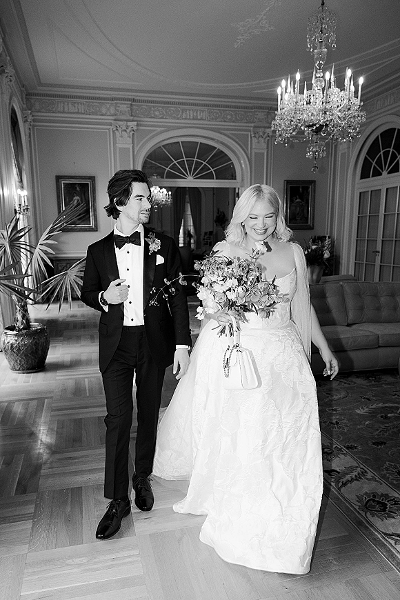 Couple portrait in a black and white wedding portrait style, bride in strapless dress and veil holding bouquet beneath a chandelier in an ornate ballroom