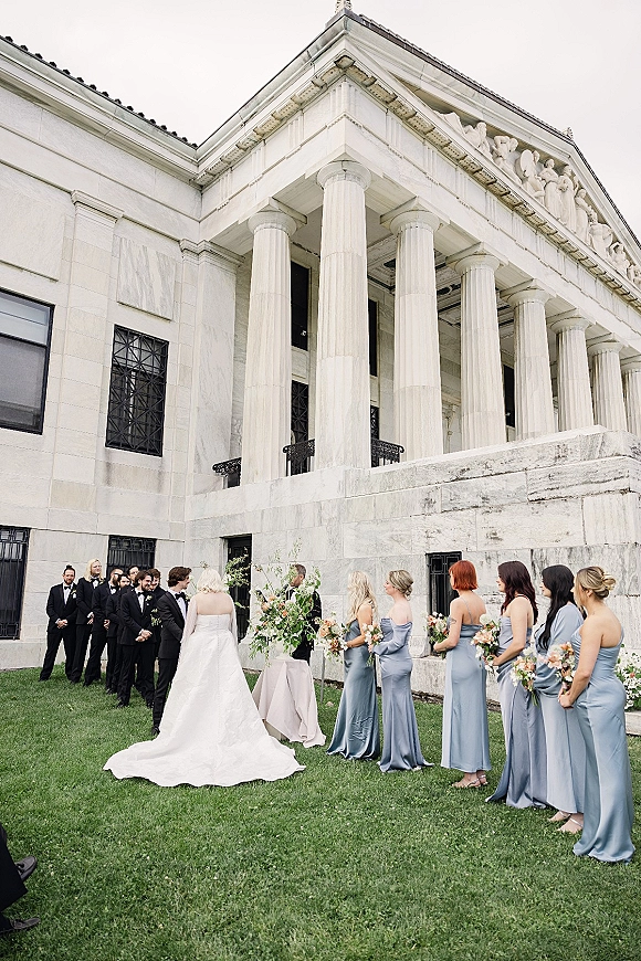 Wedding ceremony with bride in a strapless gown walking toward groom as bridesmaids in blue and tuxedoed groomsmen line up by columns
