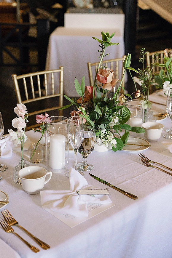 Reception tablescape with wedding table centerpiece of roses and greenery, pillar candles, gold-rim china, gold flatware at an indoor head table