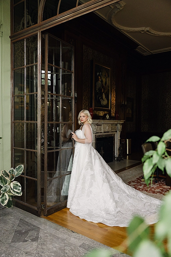 Bridal portrait of a bride in a strapless wedding dress with a long train and veil, posed by glass doors in a vintage interior
