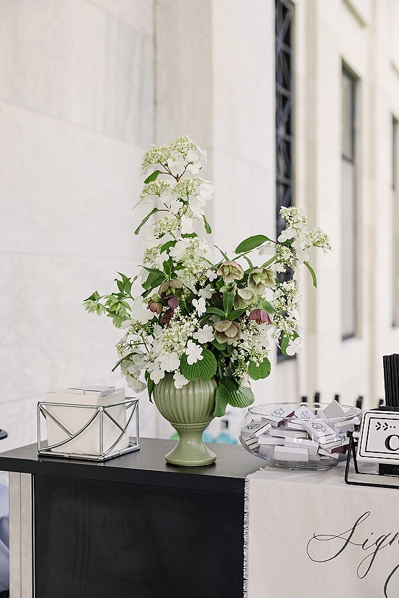 Wedding welcome table with wedding sign in calligraphy, green vase florals, favor boxes, and glass bowl on an outdoor walkway by a white building