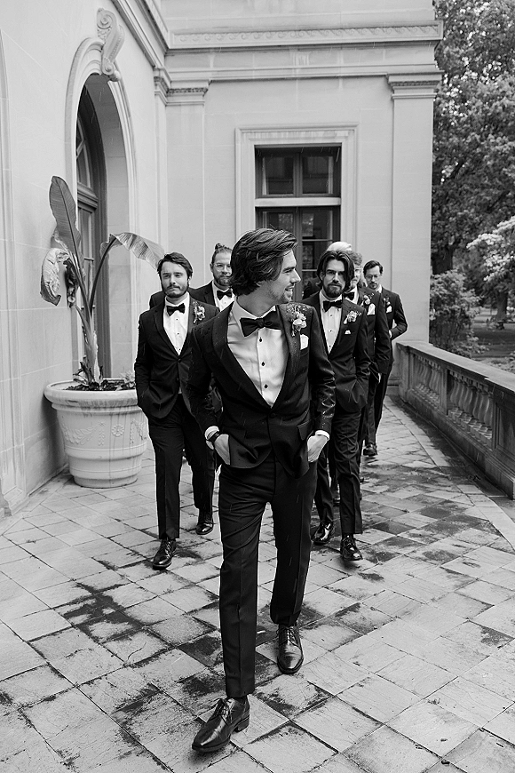 Groomsmen portrait in black tuxedos with bow ties and boutonnieres lined up on a stone terrace by an arched doorway