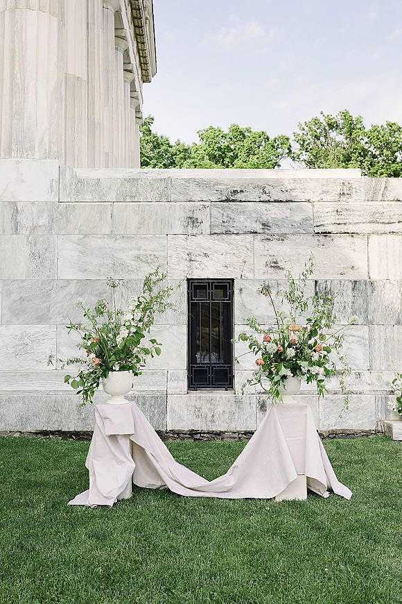 Wedding ceremony decor with ceremony entrance florals in white urns on pedestal stands, draped fabric and peach-white blooms on a lawn by a stone wall