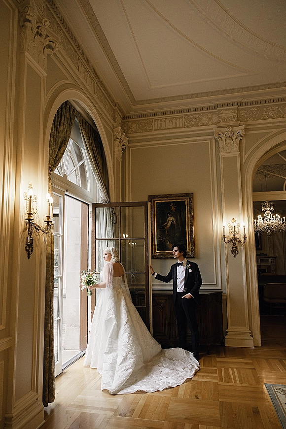 Couple portrait of bride and groom indoors, bride with bouquet and long train by arched doorway under chandelier in ornate hall