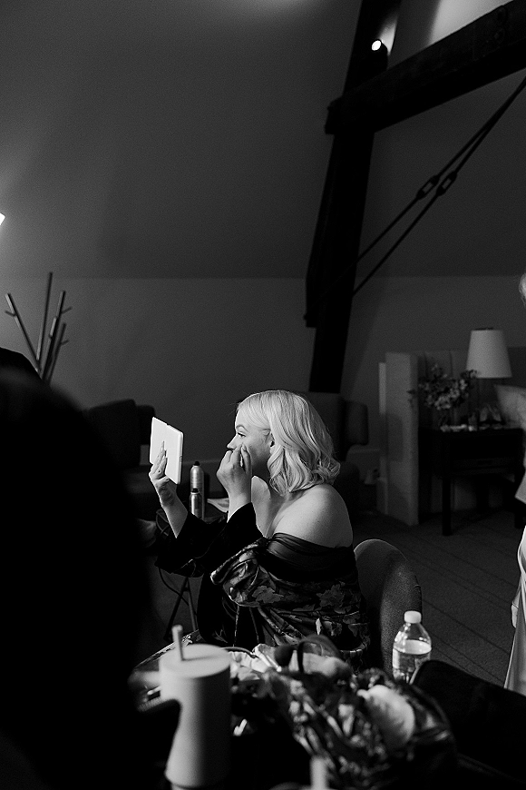 Bridal makeup moment as the bride applies lipstick while holding a hand mirror in her robe, seated in a room with exposed beams