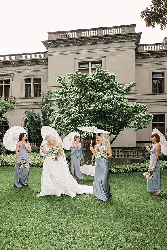 Bride with bridesmaids in blue satin dresses holding white paper parasols and bouquets on a manor house lawn with veil flowing