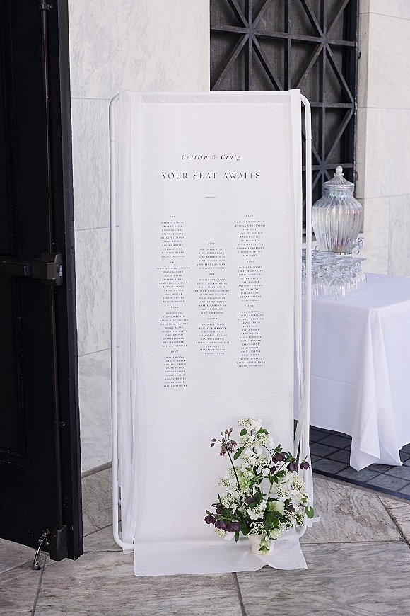 Wedding seating chart seating chart sign on a metal stand with white fabric drape and floral arrangement by a marble wall and black door