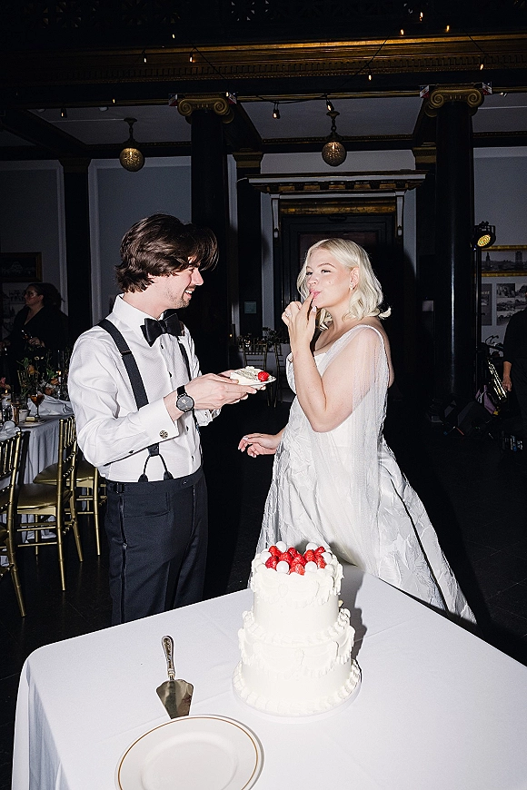 Cake cutting moment as bride in a gown and groom in suspenders slice a strawberry-topped cake under bistro string lights at an indoor reception