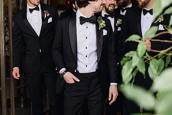 Groomsmen portrait in black tie tuxedos with satin lapels and boutonnieres, standing in a doorway framed by greenery