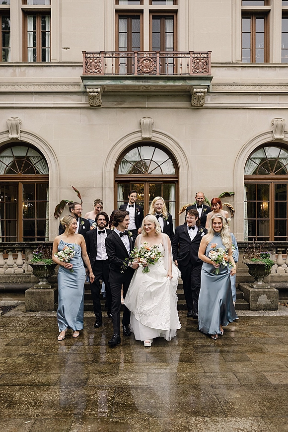 Wedding party portrait with bride and groom with wedding party walking down stone steps by arched windows on wet pavement, bouquets in hand