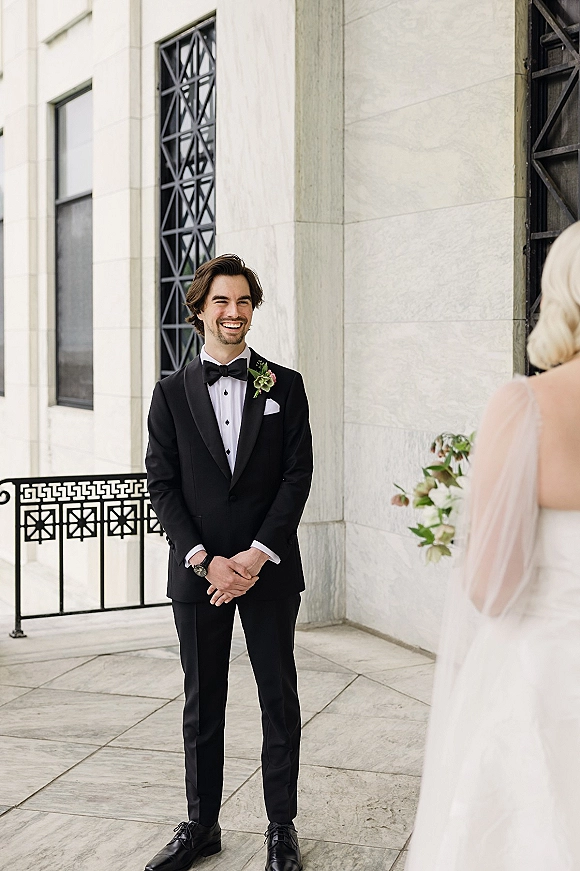 Groom portrait in a black tuxedo groom look with bow tie and boutonniere, hands clasped on a stone terrace by large windows