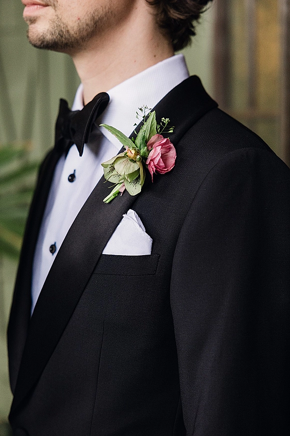 Groom boutonniere with a pink boutonniere flower and greenery pinned to a black tuxedo lapel, bow tie and pocket square, outdoors blurred greenery