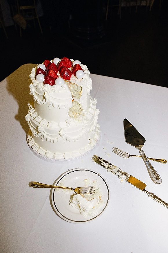 Wedding cake with two tier wedding cake design, white buttercream frosting, strawberry topping and sugar pearls on a stand with gold flatware