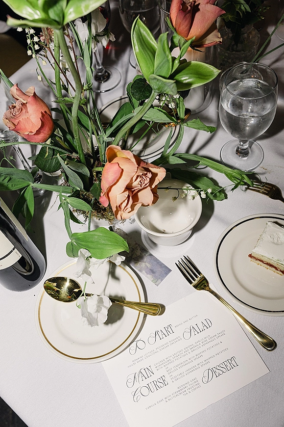 Reception tablescape wedding table setting with peach rose centerpiece, greenery, gold flatware, menus, wine glasses, and cake slice on plate