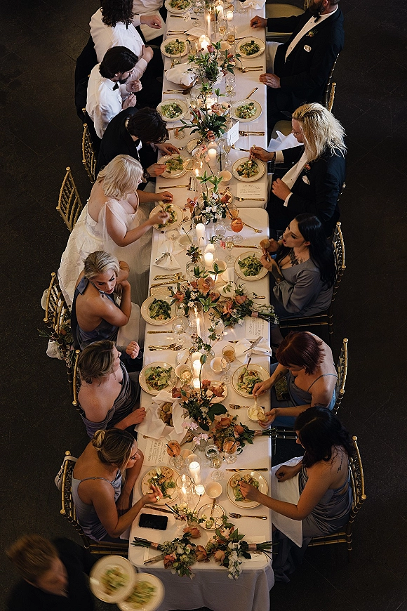 Reception tablescape on a long banquet table with floral garland, peach blooms and taper candles, set with gold flatware in an indoor space