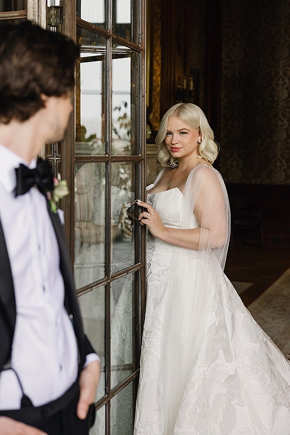 Bridal portrait of a bride in a strapless wedding dress and veil holding a glass door handle, window light on patterned wallpaper behind
