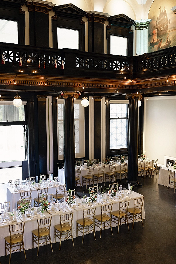 Reception tablescape with long banquet table setup, white linens, gold Chiavari chairs, bud vases, and string lights in an airy loft hall