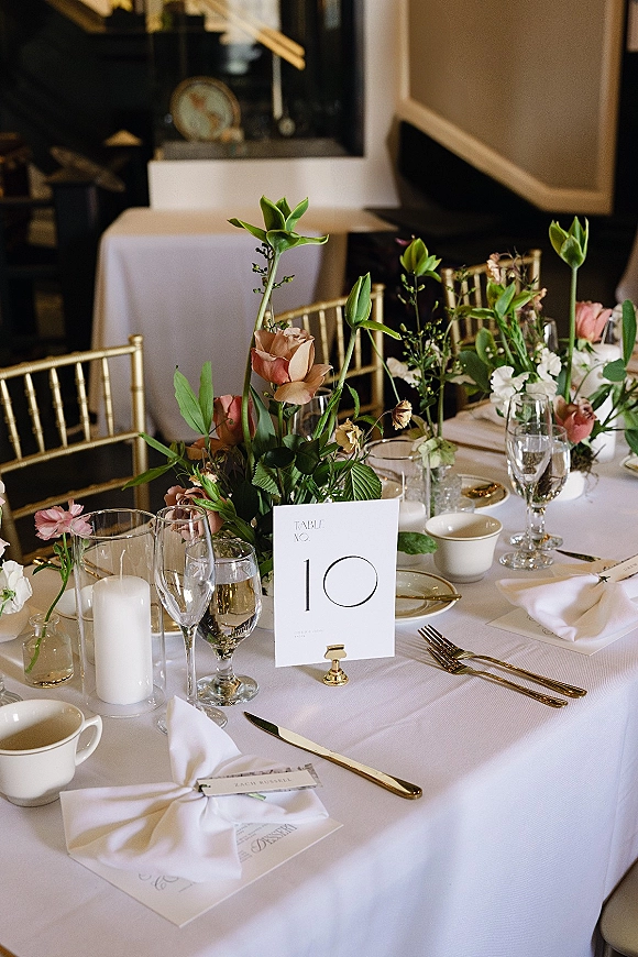Reception tablescape with a wedding table centerpiece of blush flowers and greenery, gold flatware, menu and place cards, and candles in an indoor room