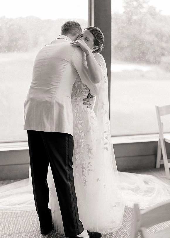 First dance as bride in lace wedding dress hugs groom in a light suit, long veil trailing by large reception windows, intimate moment