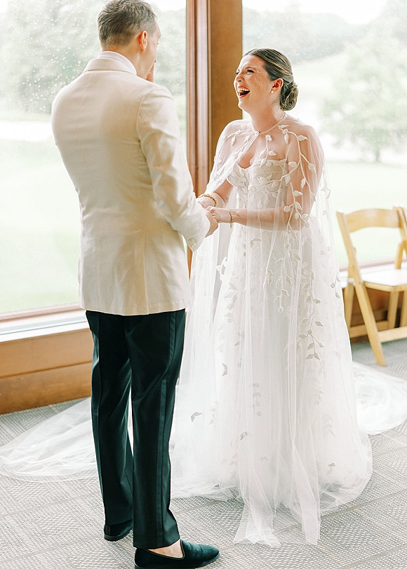 First look moment as bride in wedding dress laughs holding hands with groom in cream jacket by large windows and greenery, veil trailing