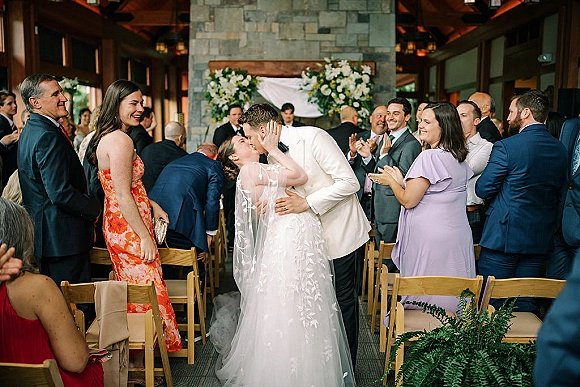 Ceremony kiss as bride in veil and gown dips with groom in white tux under a floral arch, guests cheering in rustic lodge space