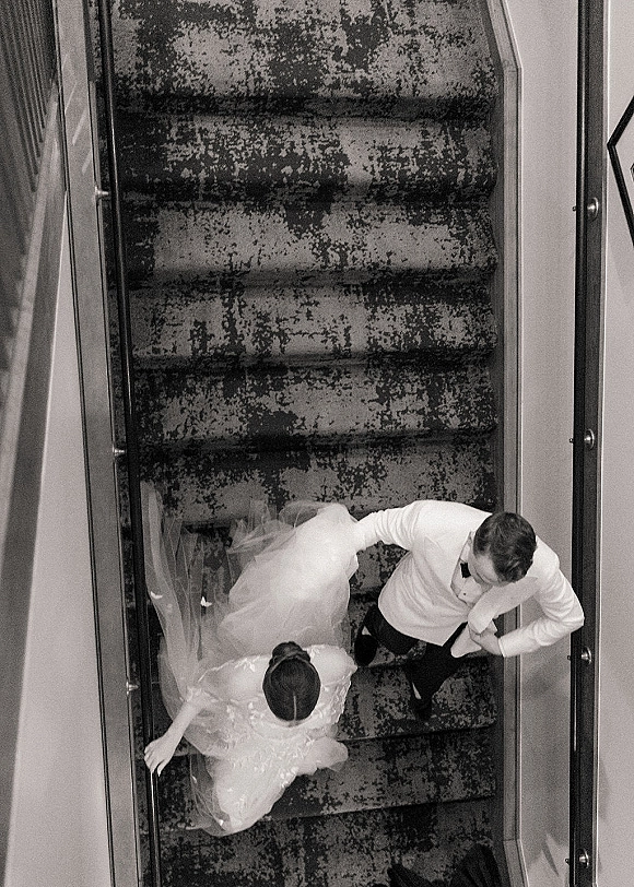 Couple portrait from above of bride and groom holding hands on a staircase, her veil and dress train flowing behind him in a tuxedo