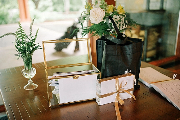 Wedding guest book table with a glass card box, envelopes and greeting cards beside a floral arrangement on a wood table in window light