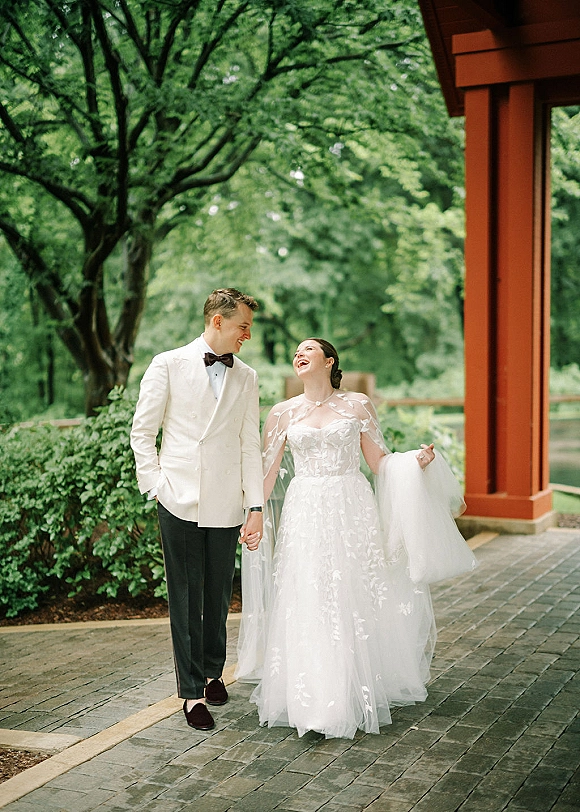 Wedding couple portrait of bride and groom walking hand in hand, bride laughing in floral gown with sheer cape, garden path behind them