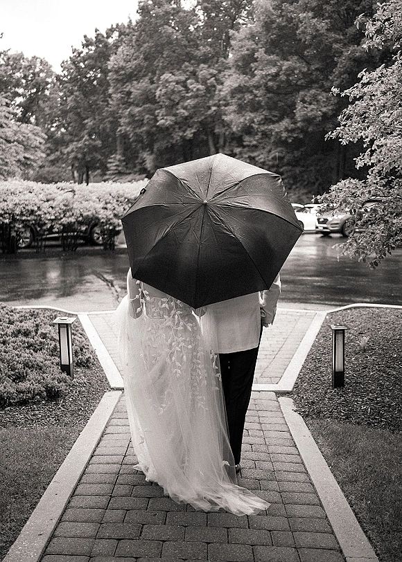Couple portrait of bride and groom under umbrella, walking away on a rainy brick path with veil trailing and pathway lights glowing