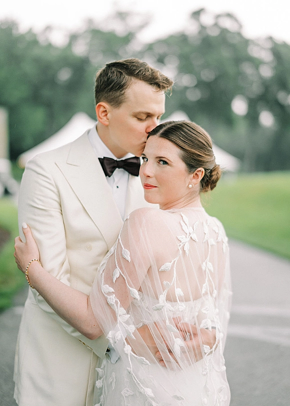 Couple portrait with groom kissing bride’s forehead as she looks over her shoulder, wearing a sheer embroidered cape on an outdoor lawn by trees and a white tent