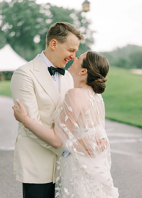 Couple portrait of bride and groom embrace, smiling with foreheads touching, her lace veil draped as he wears a white tux on an overcast lawn