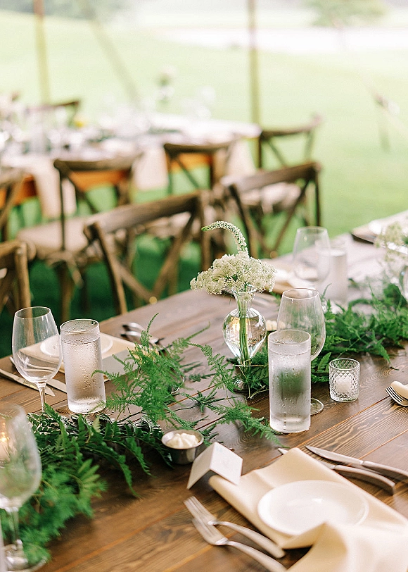 Reception tablescape with rustic wedding table setting, greenery garland runner, bud vases, votive candles, and crossback chairs under a tent