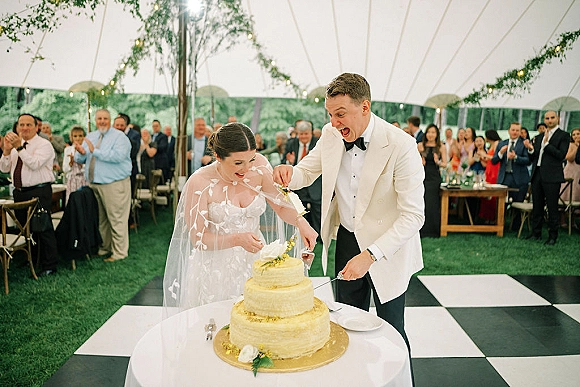 Wedding cake cutting as bride in veil and groom in white dinner jacket slice a tiered cake under bistro lights in a tent reception